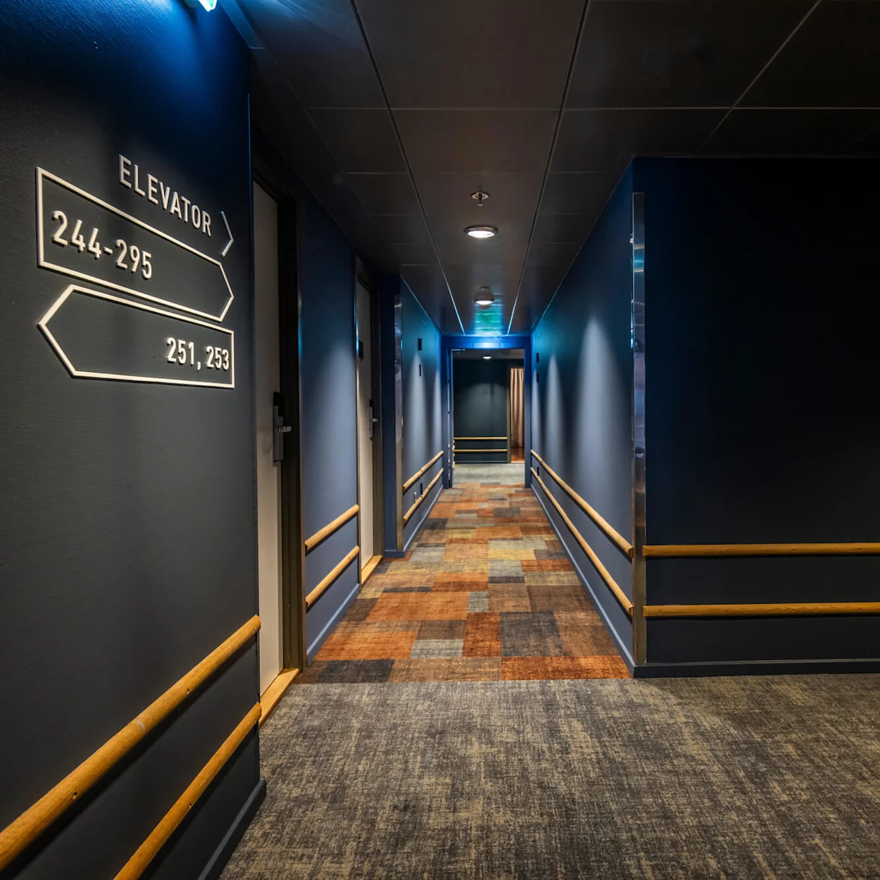 A hotel corridor with dark blue walls, wooden rails along the walls, and a carpet pattern in warm tones. A sign on the wall indicates room numbers and the direction to the elevator.