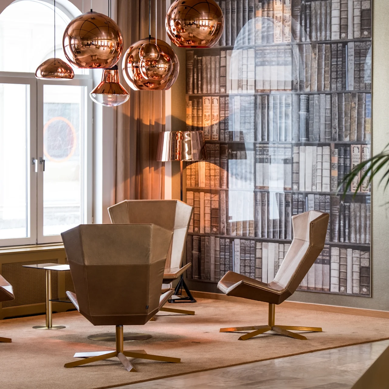 Fire moderne stole omgiver et lille bord under kobberlamper i et rum med en væg designet som bogreoler. Store vinduer og planter skaber en indbydende atmosfære.