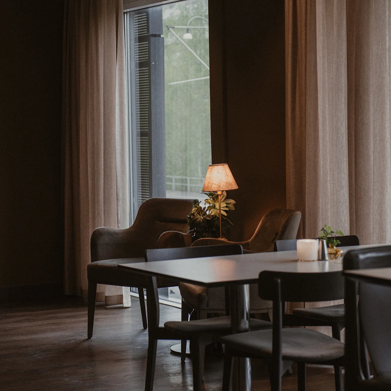 A cozy armchair sits by a window, softly illuminated by a lamp on a side table, in a dimly lit room with surrounding dining tables and chairs.