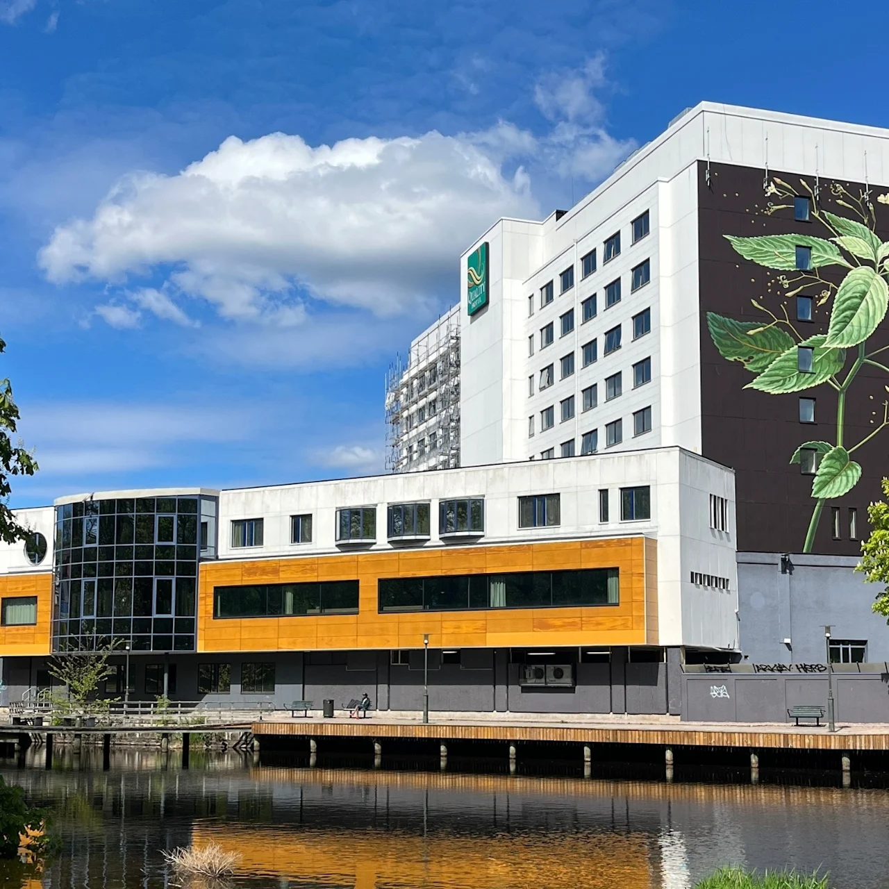 A multi-story building displays a large botanical mural on its facade. It stands beside a calm waterway, surrounded by lush green trees, under a clear blue sky. Text: "SCANDIC".