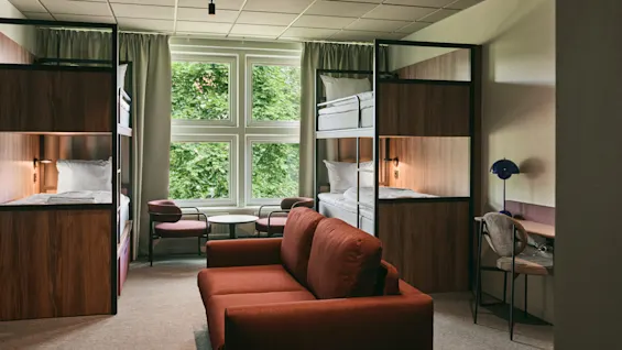 A modern shared room with bunk beds, a comfortable red sofa, and a desk, bathed in natural light from a large window.