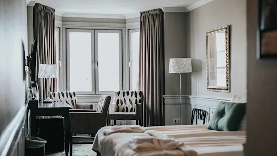 Elegant møbleret soveværelse med redt seng, stole, skrivebord og lamper, der formidler en hyggelig, veloplyst atmosfære.