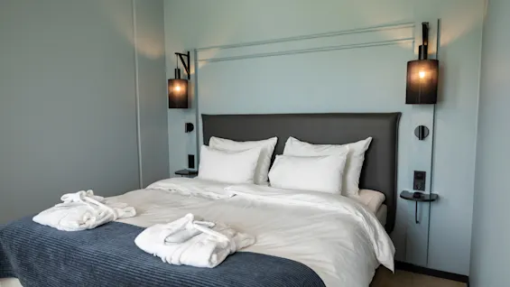 Et koselig hotellrom med en stor seng, hvitt sengetøy, et blått pledd og to badekåper med tøfler.
