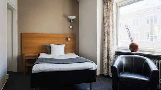 En enkelsäng med vita lakan och grå filt står mot en brun vägg i ett hotellrum, bredvid ett fönster med mönstrade gardiner och en svart fåtölj.