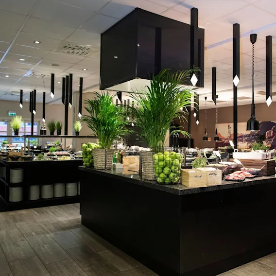 A spacious buffet area features counters laden with fresh greens, fruits, and various foods, all under stylish pendant lights. Green plants and baskets filled with apples add vibrant accents. Text on the wall reads: “GLUTEN-FREE LACTOSE-FREE”.
