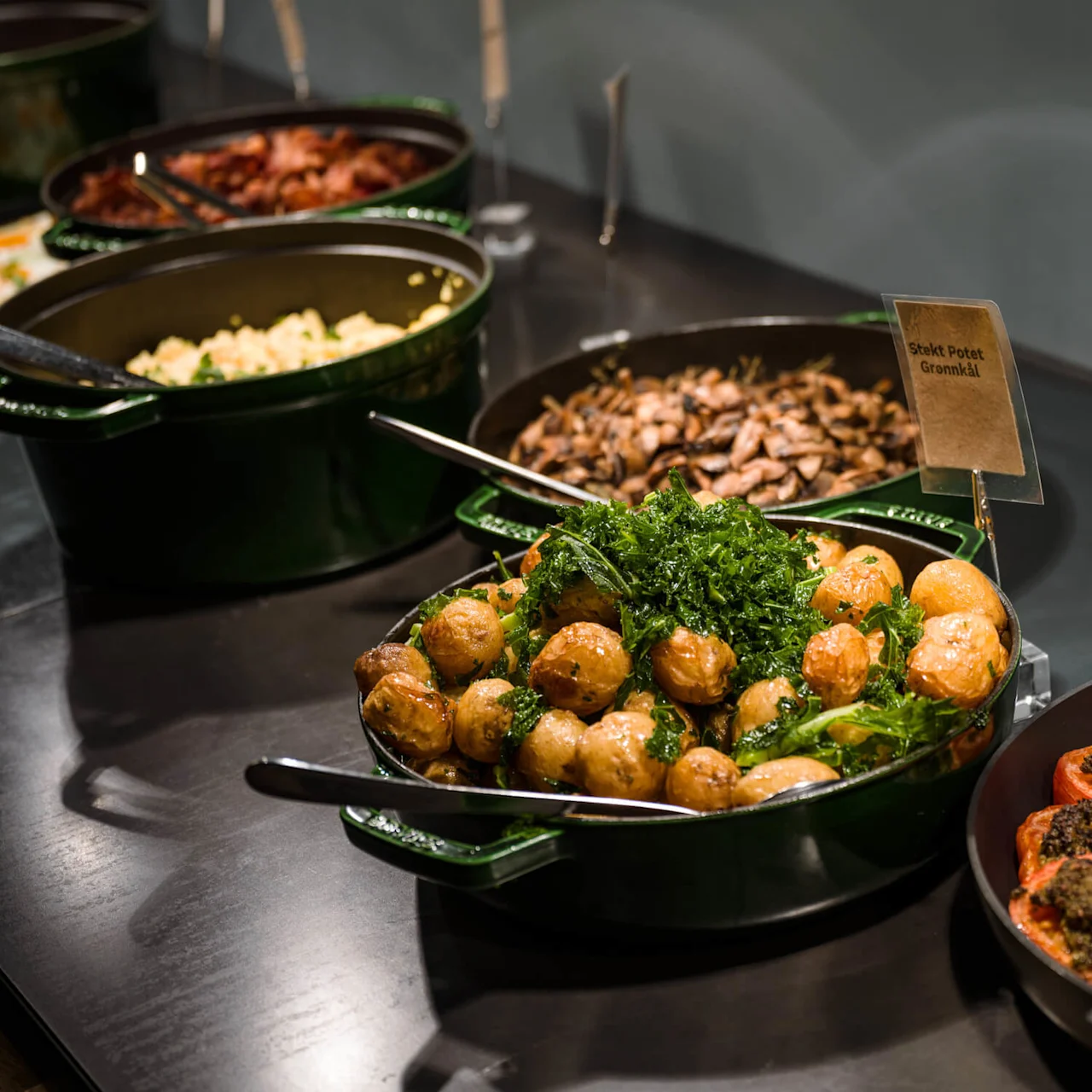 Stekte poteter med grønnkål vist i en grønn serveringsskål, sammen med stekte sopp og ristede tomater på en buffébord. Skiltet sier: "Stekt Potet Grønnkål".