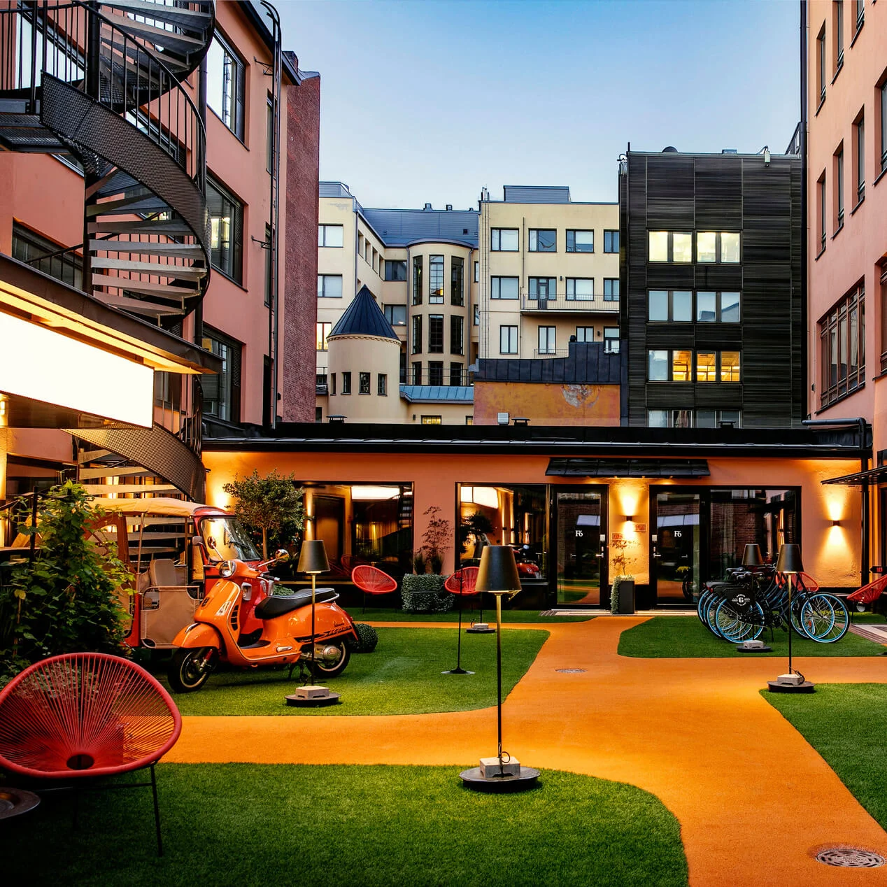 Orange scooter parkert på kunstgress med utelamper og røde stoler i en gårdsplass omgitt av moderne bygninger i flere etasjer ved skumring.