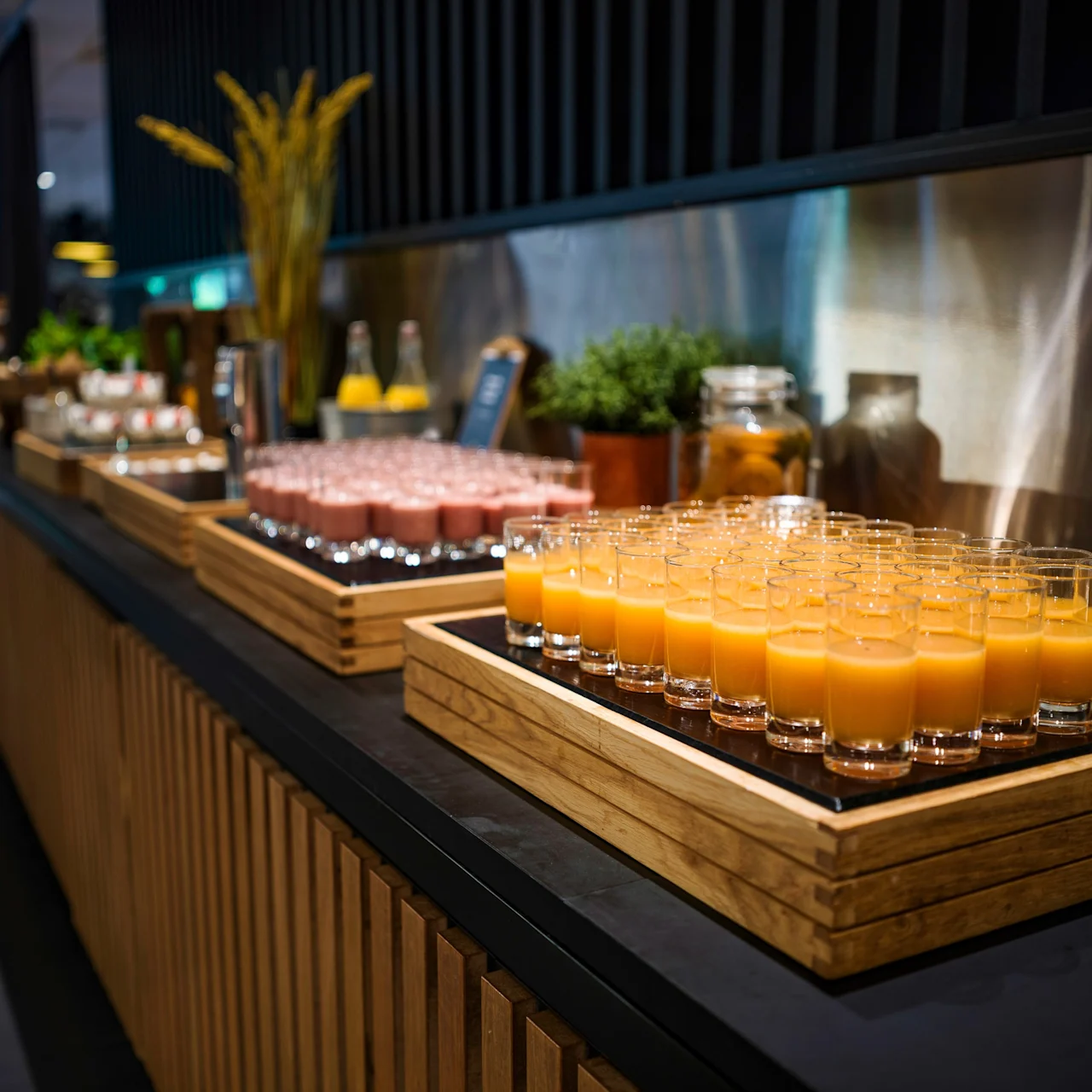 Glasses of orange juice are arranged neatly on a wooden tray on a counter. Nearby are more trays with drinks and decorative plants in a well-lit buffet setting.