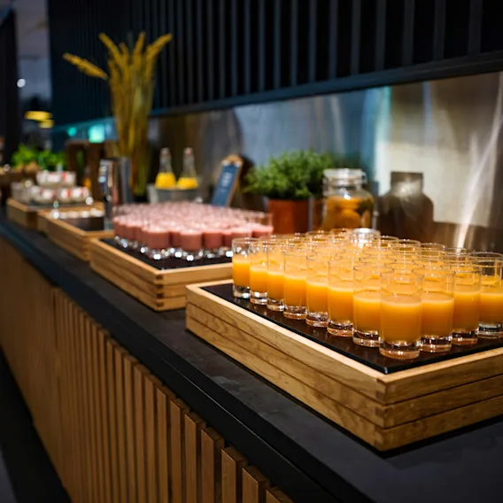 Glass med appelsinjuice er pent arrangert på et trefat på en disk. I nærheten er det flere fat med drikke og dekorative planter i en godt opplyst buffémiljø.