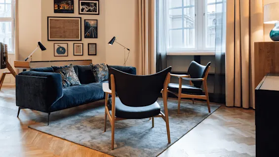 Blue velvet sofa with patterned cushions sits on a gray rug, facing two wooden chairs near large window. Framed art decorates beige walls; lamps add soft lighting.
