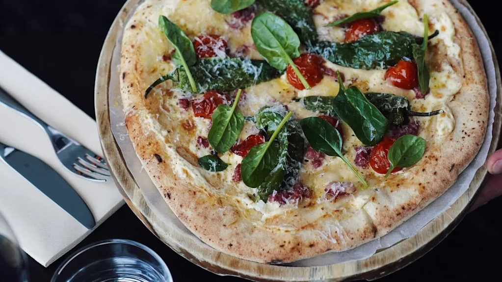 En pizza toppet med spinat, cherrytomater og ost holdes av en tatovert arm i hvit uniform. I nærheten er gaffel, kniv og glass.