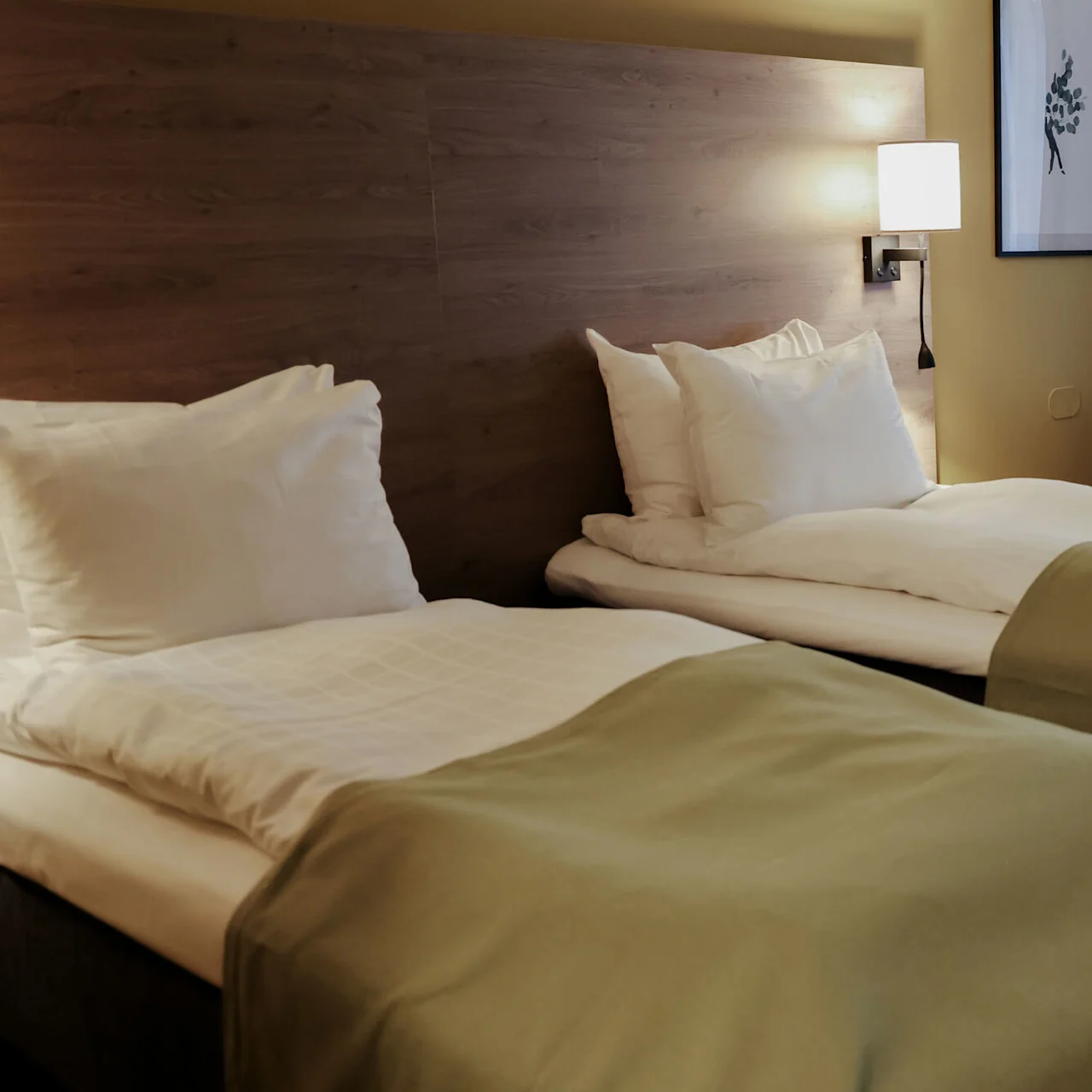 Two neatly made twin beds rest against a wooden headboard, covered with green blankets. A wall lamp illuminates an adjacent chair and curtained window, creating a cozy hotel room atmosphere.