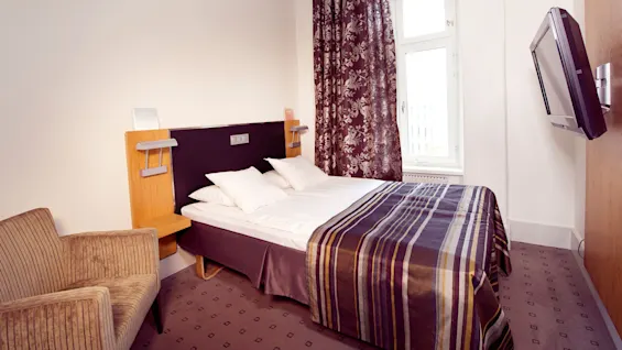 A neatly made bed in a hotel room with a striped blanket, flanked by a chair and a television on the wall.