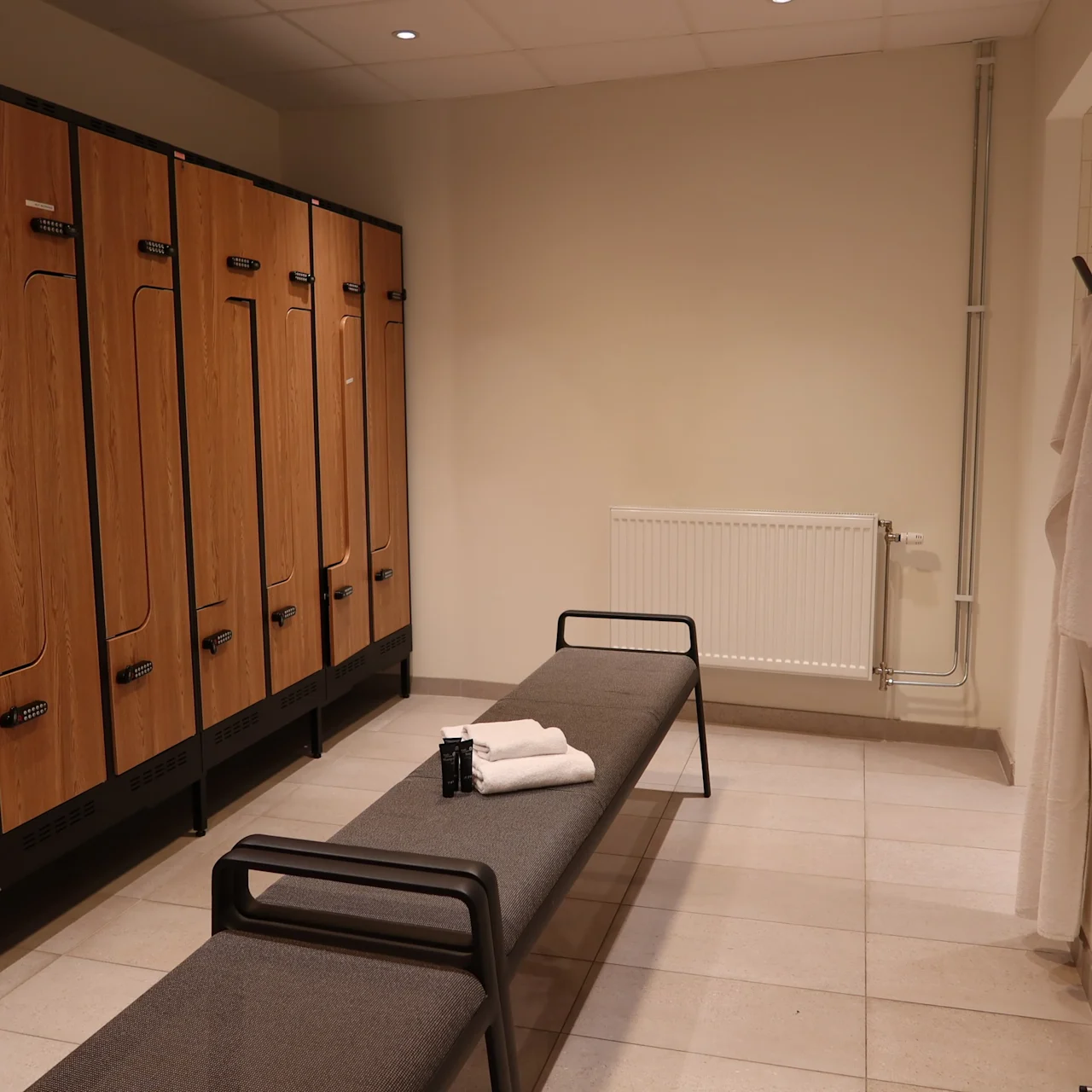 Locker room with wooden lockers along the left wall, a bench in the center holding folded towels and a toiletry bag, and a towel hanging on a hook to the right.