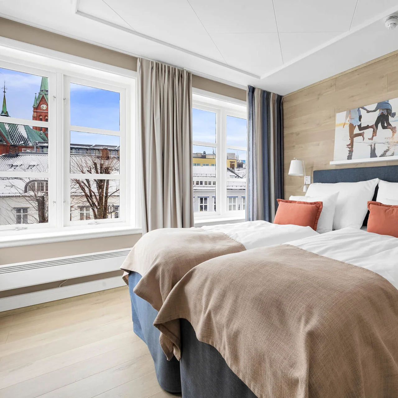 En pent seng med oransje og beige puter, vegglamper og kunst, er plassert nær store vinduer med utsikt over snødekte hustak med et kirketårn utenfor.