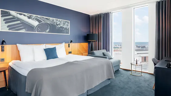 En seng med grått og hvitt sengetøy står i et moderne hotellrom. Ved siden av er en sofa og et lite bord med glass som har utsikt over byen gjennom store vinduer.
