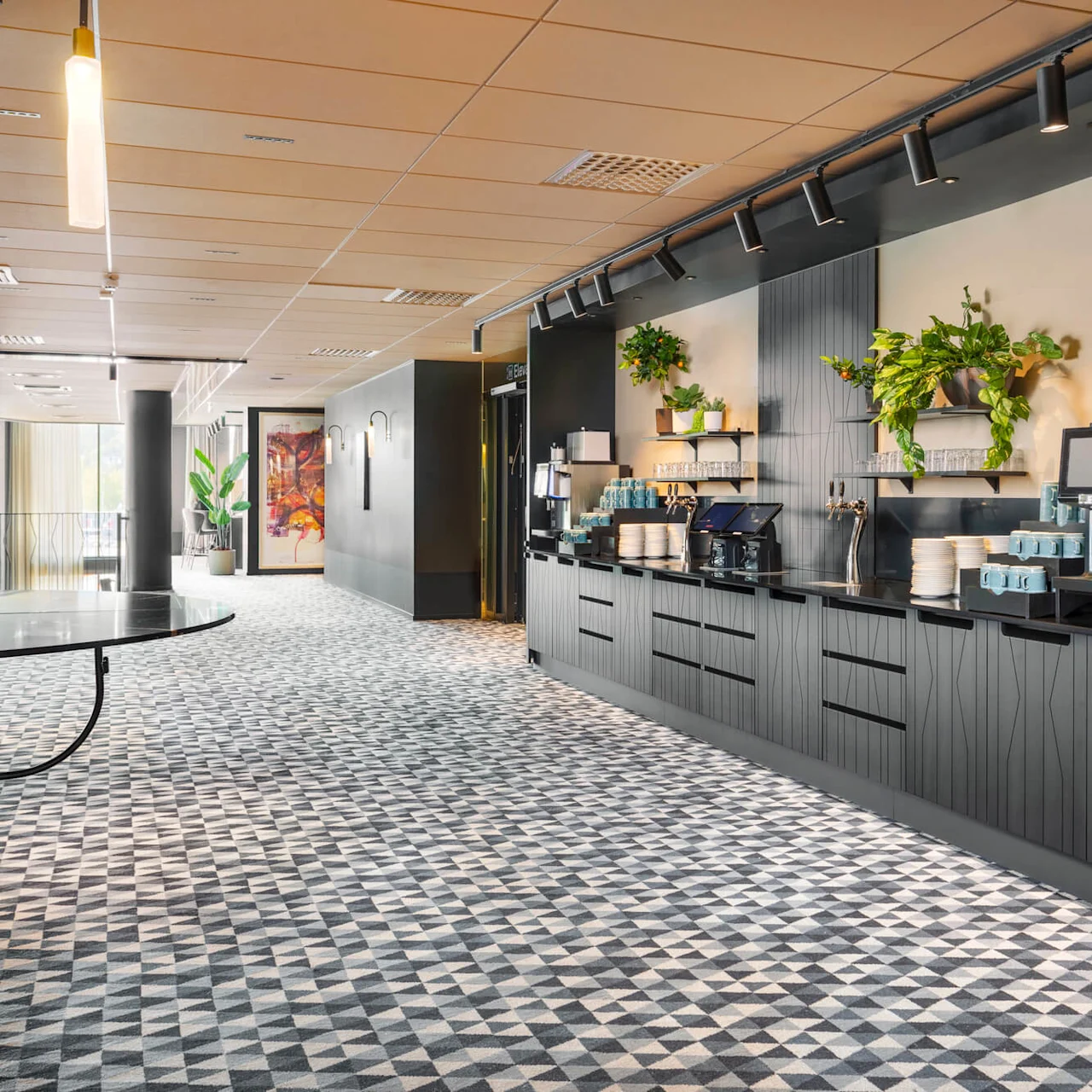 Kaffestation med modern design, innehållande kaffemaskiner och koppar på en mörk bänk. Geometriskt mönstrad matta och gröna växter förhöjer den moderna atmosfären i den öppna gången.