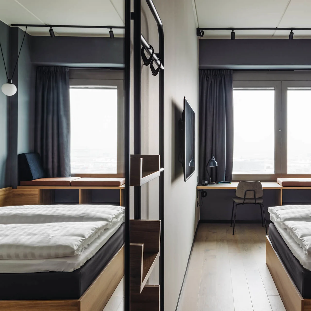 A modern hotel room features two beds separated by a partition, with a desk and chair in front of large windows, and dark curtains providing a cozy ambiance.