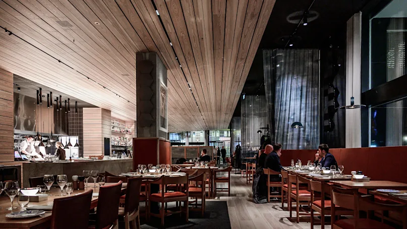 A modern restaurant with wooden interiors features people dining at tables with wine glasses. Waitstaff prepare meals at an open kitchen, contributing to an elegant, lively atmosphere.