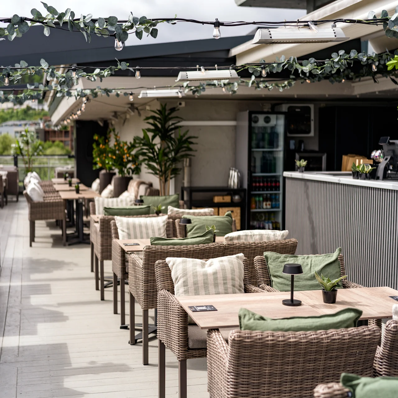 Café tables with wicker chairs are arranged on a rooftop terrace. String lights and plants create a cozy ambience in a modern setting with a view of a distant landscape.