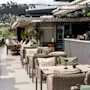 Café tables with wicker chairs are arranged on a rooftop terrace. String lights and plants create a cozy ambience in a modern setting with a view of a distant landscape.