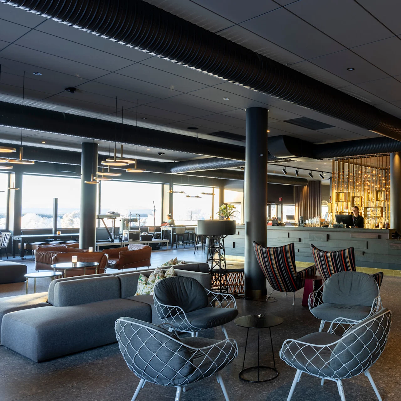 En modern lobby har stiliga stolar och soffor i ett rymligt område. En personal står vid en elegant reception, omgiven av dekorativ belysning. Stora fönster ger naturligt ljus.