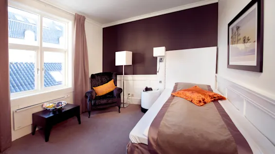 En prydligt bäddad säng dominerar ett välupplyst hotellrum, flankerad av dekorativa lampor och en plyschfåtölj, med utsikt över tegeltak.