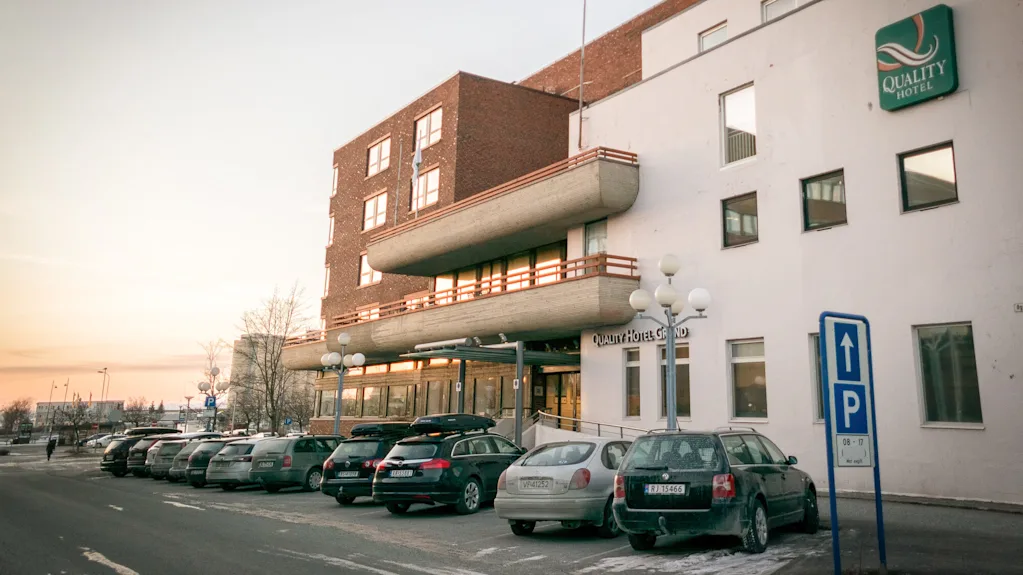 Et hotel med et "Quality Hotel" skilt på facaden, biler parkeret foran og et blåt P-skilt. Himlen er blødt oplyst, hvilket antyder solopgang eller solnedgang.
