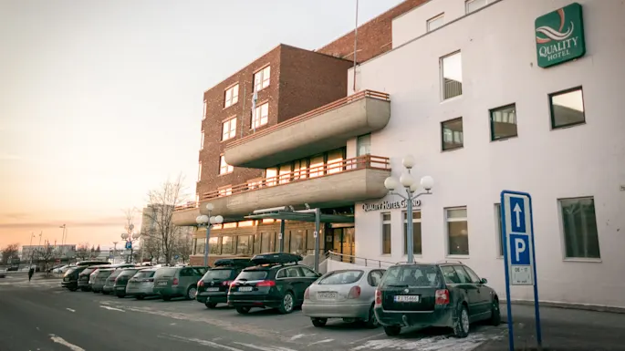 A hotel with a "Quality Hotel" sign on the facade, cars parked in front, and a blue parking sign. The sky is softly lit, suggesting sunrise or sunset.