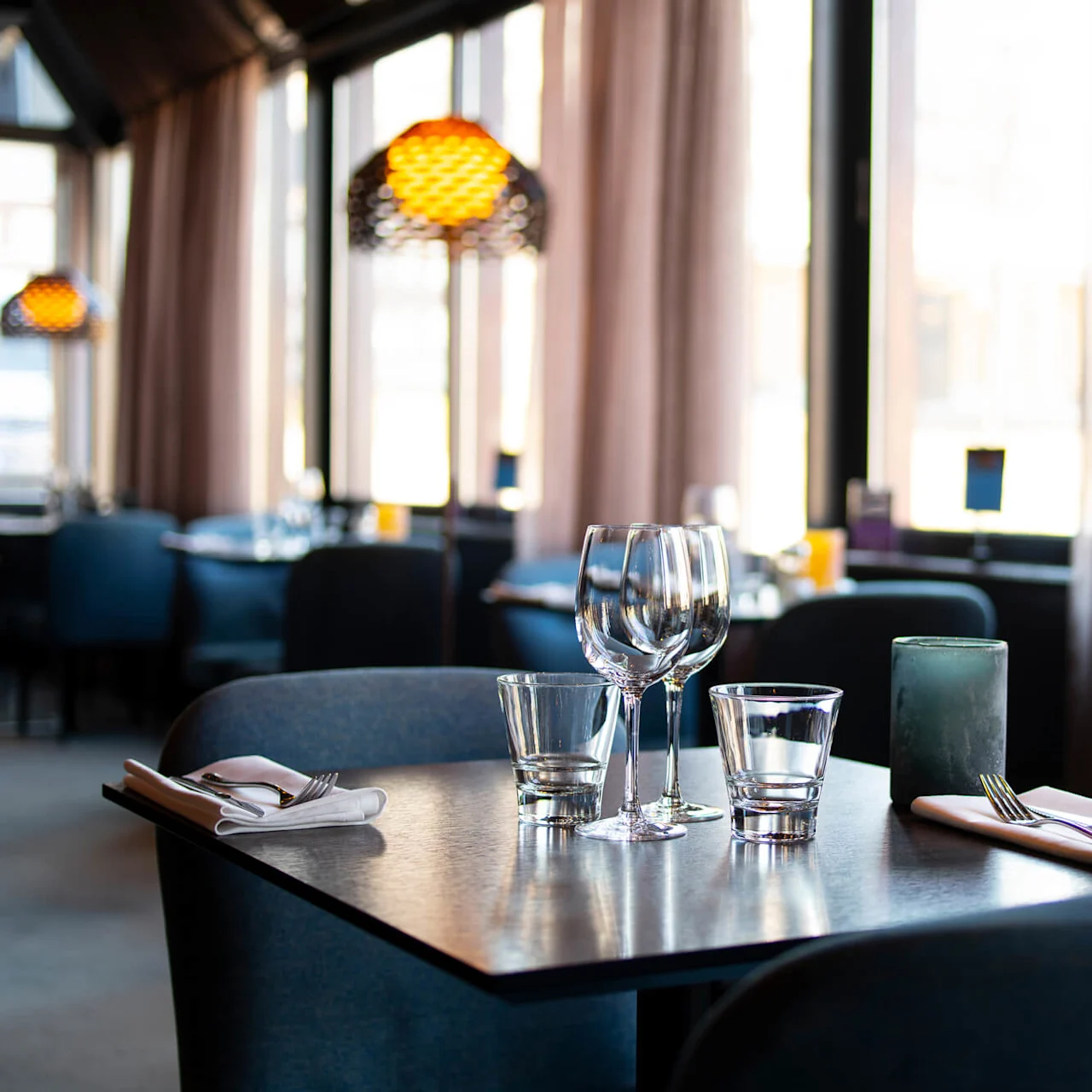 Ett bord dukat med glas och bestick i en svagt upplyst restaurang, omgivet av blå stolar och stora fönster med gardiner.