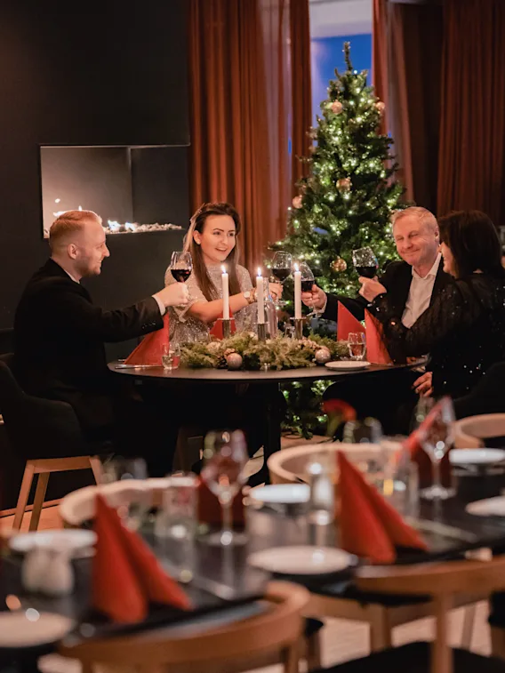 Fire personer hever vinglass for en skål, sittende rundt et bord med stearinlys og grønt, foran et dekorert juletre.