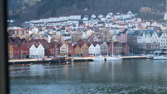 En livlig by ved vannet med fargerike bygninger, båter fortøyd i havnen og en frodig grønn åsside i bakgrunnen.