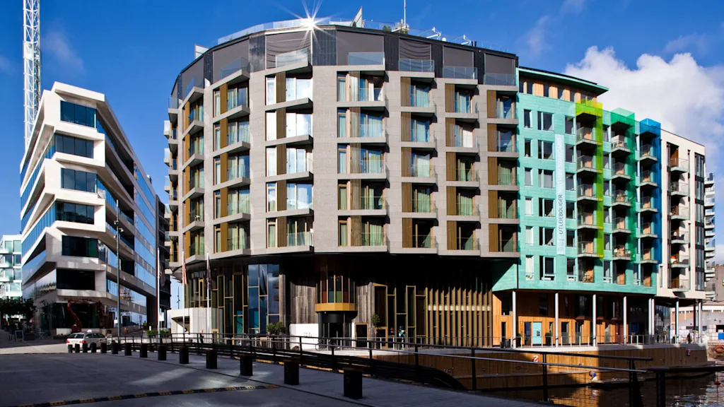 A modern, angular building with glass balconies and multicolored sections stands against a clear blue sky. It's situated by a waterfront, reflecting contemporary architectural design.