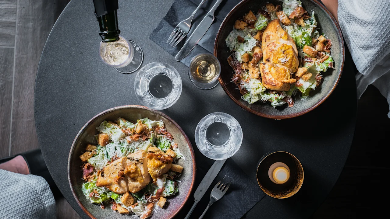 Två läckra kyckling Caesar-sallader med drycker och ett ljus på ett mörkt bord, redo för en mysig måltid.