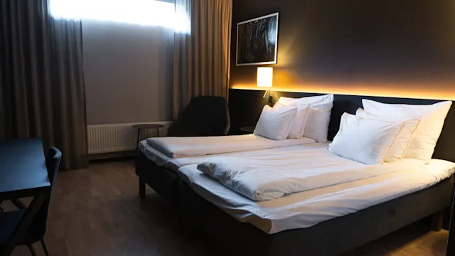 Huolellisesti pedattu sänky valkoisine liinavaatteineen on keskellä, vierellään pehmeä valo tummalla seinällä. Lähellä verhot peittävät ikkunan, päästäen pehmeää valoa huoneeseen.