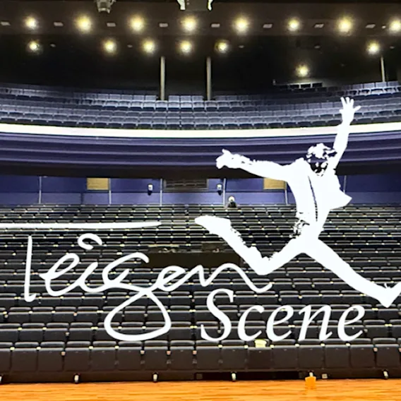En person hopper på en scene i et stort auditorium med rader av seter. Ordene Teigen Scene vises.