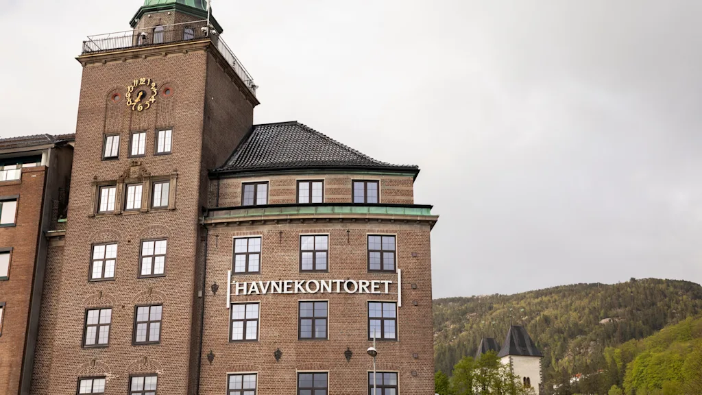 En stor murbygning med et klokketårn viser teksten "HAVNEKONTORET" på fasaden. Det er omgitt av skogkledde åser og en overskyet himmel.