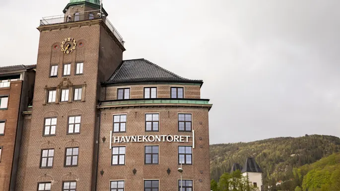 A large brick building with a clock tower displays the text "HAVNEKONTORET" on its facade. It is set against a backdrop of wooded hills and cloudy sky.