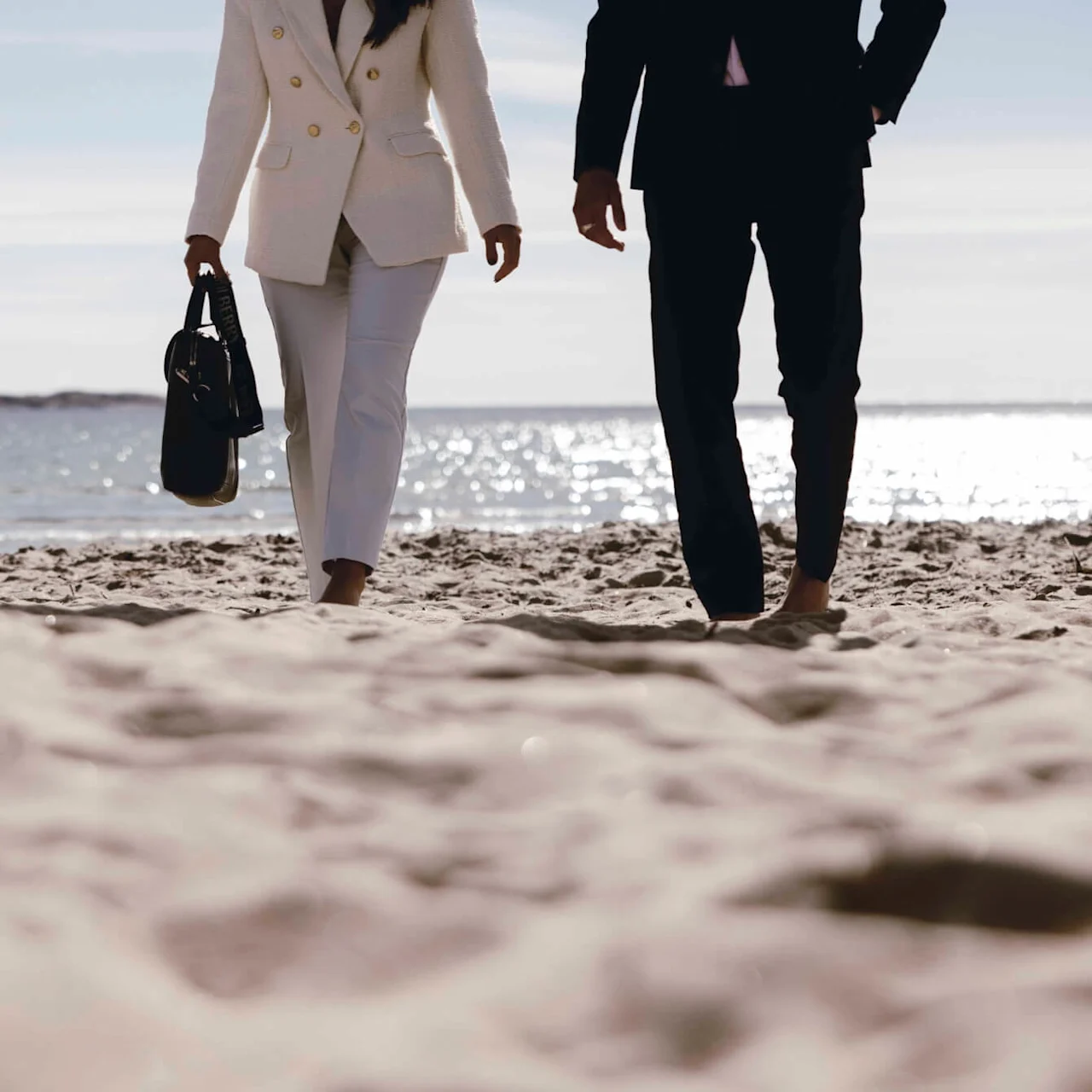 Two people in formal attire walk barefoot on a sandy beach, with the ocean sparkling in the background under a clear sky.