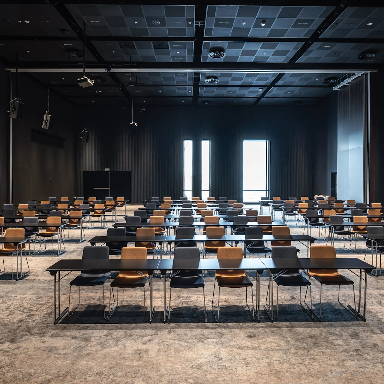 Rækker af sorte skriveborde med brune stole er arrangeret i et rummeligt, tæppebelagt konferencelokale, oplyst af høje vinduer i baggrunden.