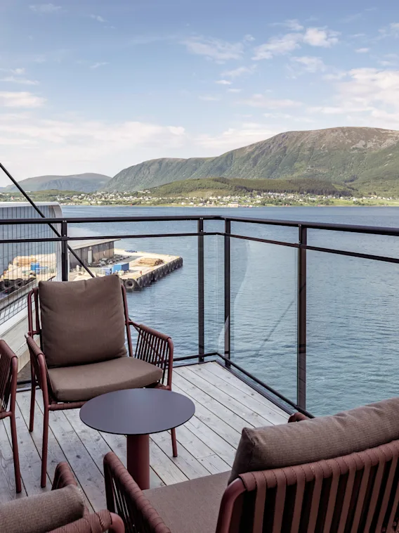 Fyra kuddförsedda stolar omger små bord på en balkong med utsikt över ett lugnt hav. Ett avlägset bergslandskap fullbordar den fridfulla miljön.