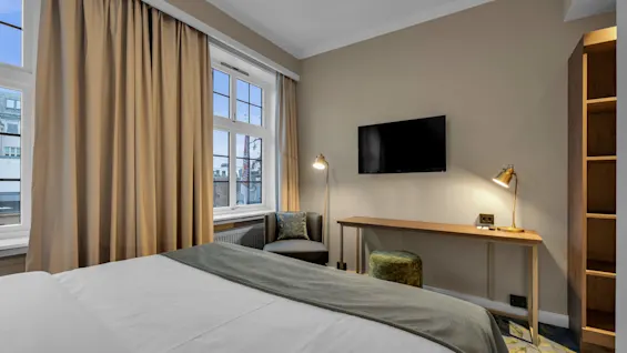 Ett snyggt bäddad säng i ett välbelyst hotellrum med ett skrivbord, stol och TV.