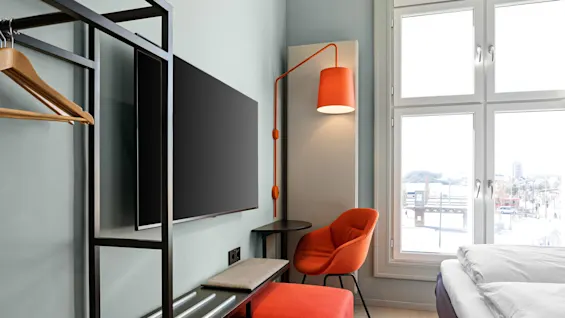TV mounted on the wall beside a modern stand, with an orange chair under a matching lamp. Large windows light the room, including a bed and minimalist furnishings.
