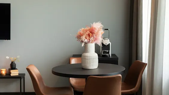 Et koselig spiseområde med et rundt svart bord, brune stoler og en vase med rosa tørkede blomster. En kaffemaskin står i bakgrunnen.