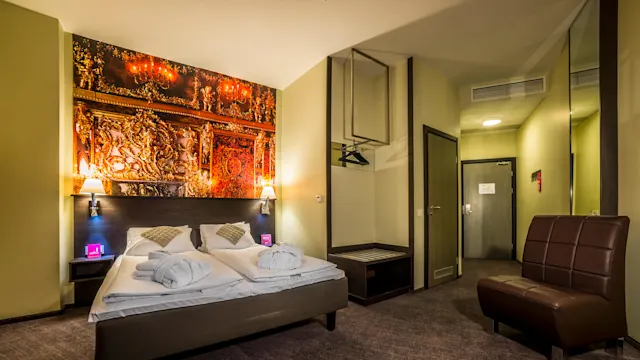 A neatly made double bed with plush pillows and folded bathrobes, positioned under an ornate wall tapestry depicting classical art, within a brightly lit hotel room with warm colors. 
