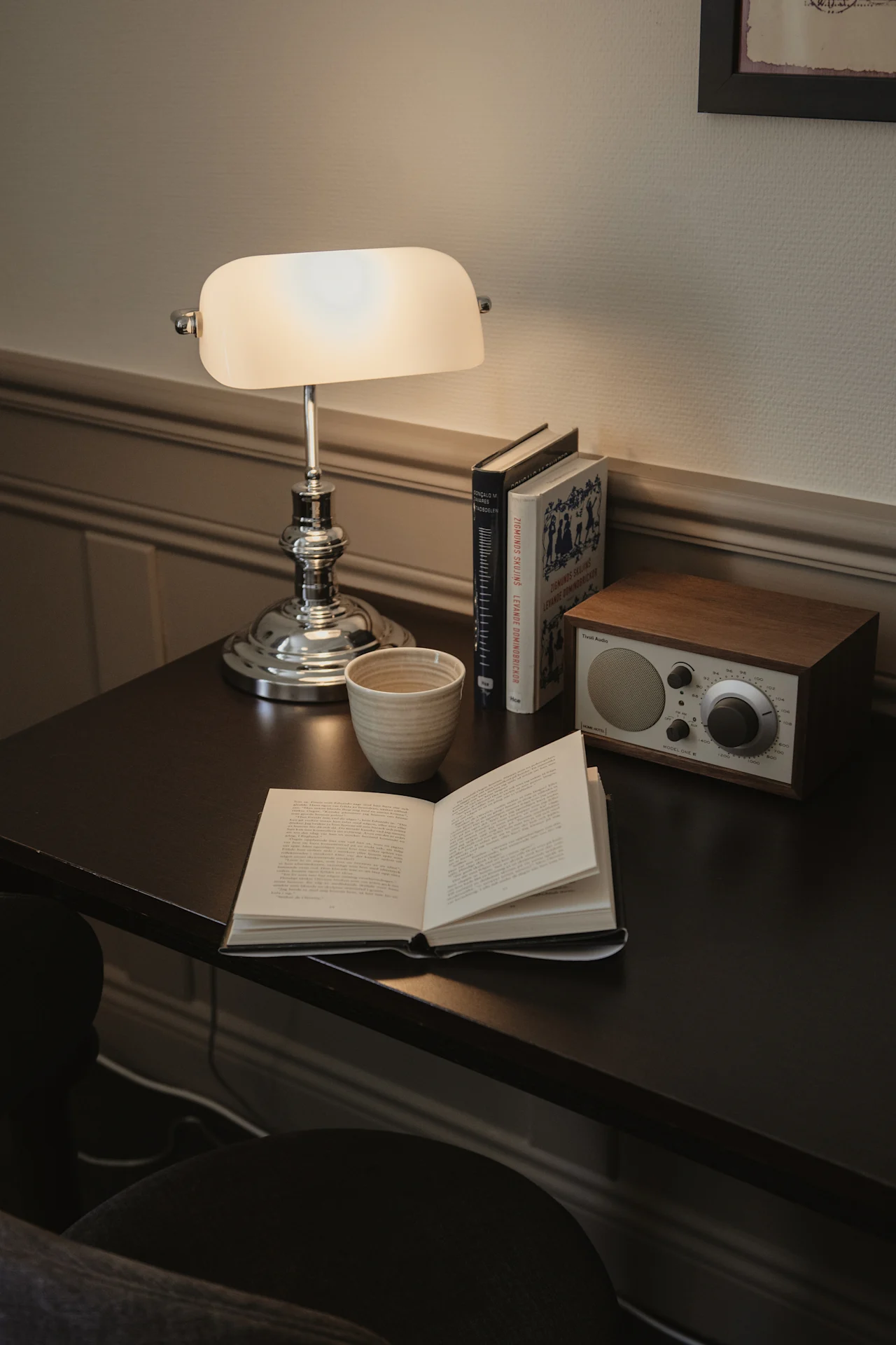 En mysig läs- eller arbetsplats med en öppen bok, en kopp och en radio på ett mörkt skrivbord, upplyst av en klassisk skrivbordslampa.
