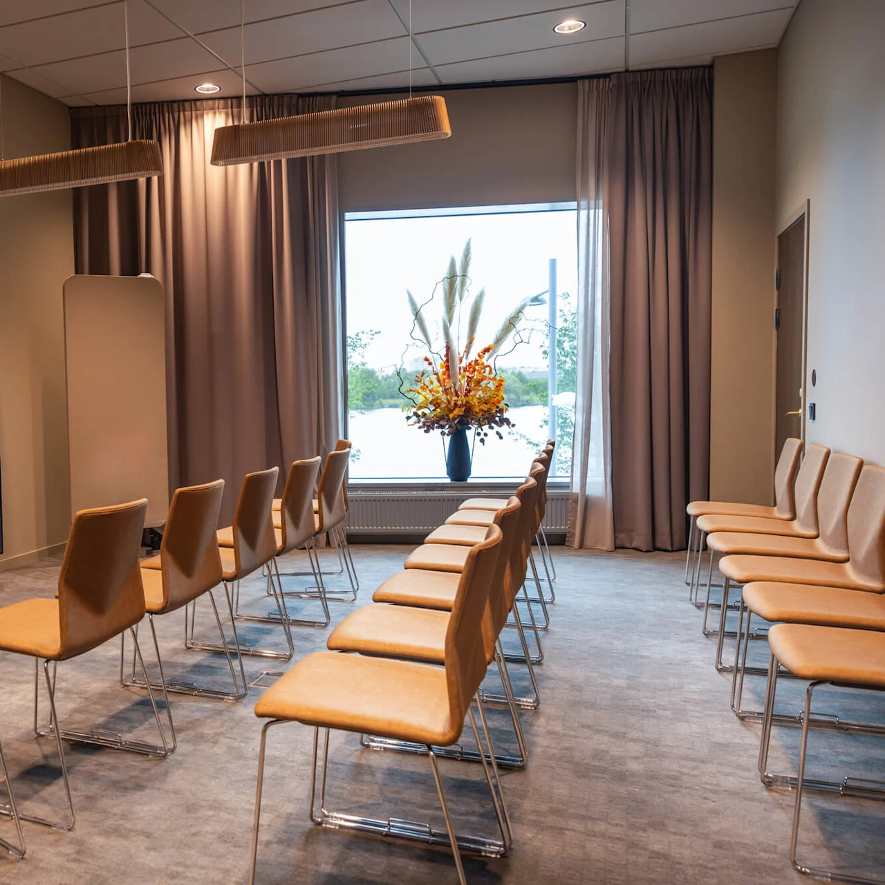 Rader av bruna stolar står vända mot en stor TV på väggen i ett modernt konferensrum, dekorerat med eleganta taklampor och ett fönster med utsikt över en naturskön vy och ett blomsterarrangemang.