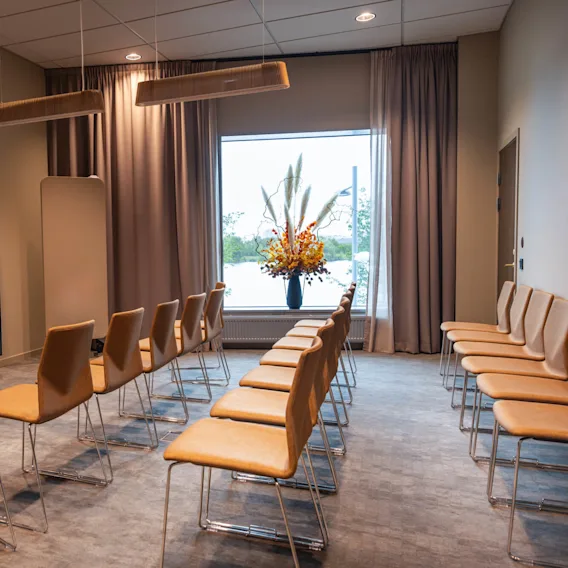 Rader med brune stoler vender mot en stor TV montert på veggen i et moderne konferanserom, dekorert med elegante taklamper og et vindu med utsikt over en naturskjønn utsikt med en blomsteroppsats.
