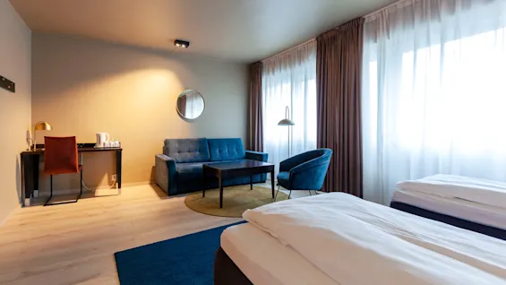 To pent oppredde senger står i et lyst hotellrom med tregulv, ledsaget av en blå sofa og lenestol, skrivebord og store vinduer med gjennomsiktige gardiner.