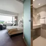 A hotel room with a neatly made double bed, visible through a partially open frosted glass door to an adjacent bathroom. The room has large windows and two gray chairs by a small table.